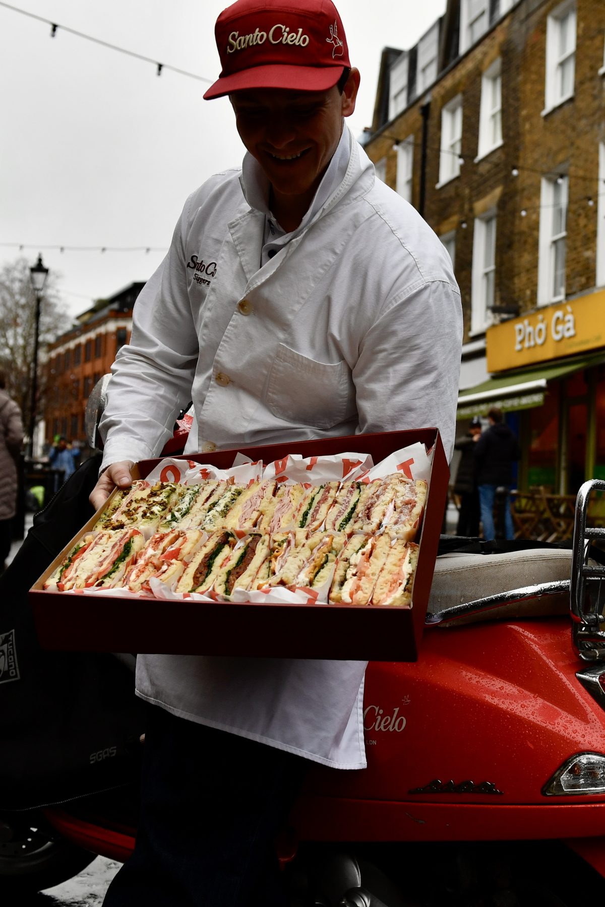A smiling Santo Cielo team member in whites and a red cap presenting an open platter box of schiacciata sandwiches on a London street