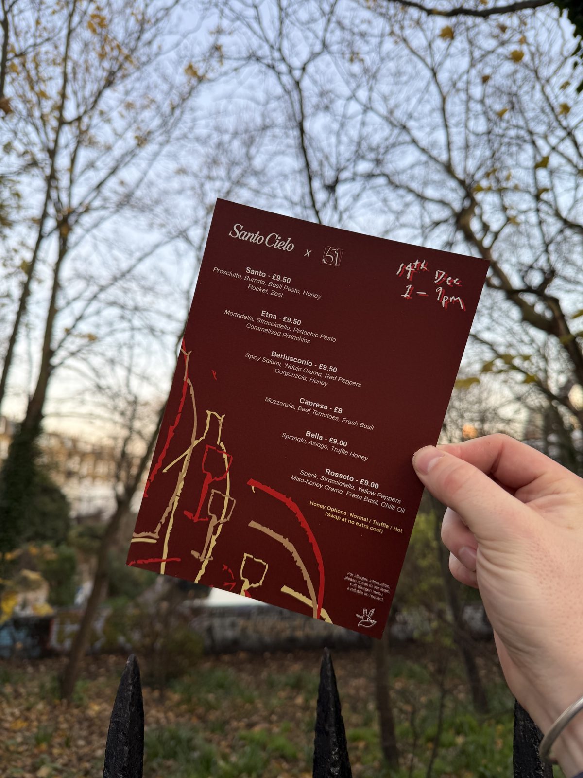 The Santo Cielo x 51 collab menu card held up outside Exmouth Market, listing Santo, Etna, Berlusconio, Caprese, Bella and Rosseto with handwritten date and times