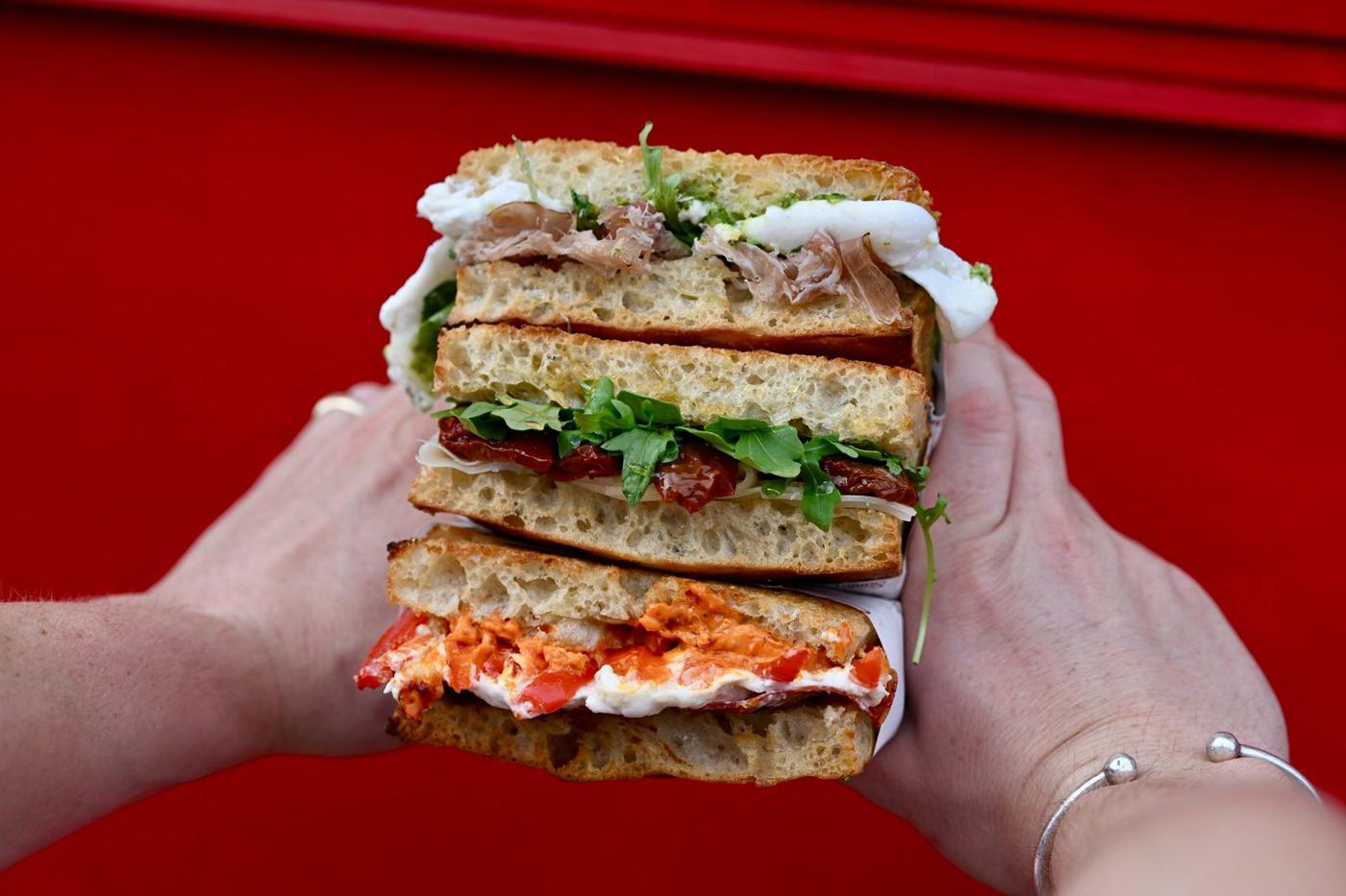 Two hands holding a stack of three cut schiacciata sandwiches against a red wall