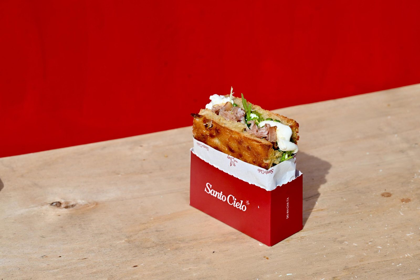 A Santo Cielo schiacciata sandwich with prosciutto, burrata and rocket standing in a red Santo Cielo box on a wooden table against a red wall