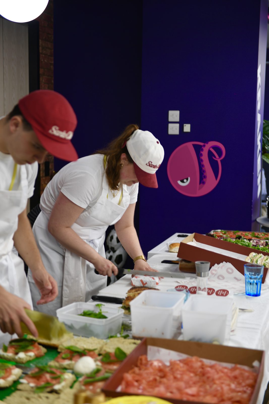 Santo Cielo team in whites and red caps building sandwiches at the Kraken Technologies office pop up, with the Kraken logo on the wall