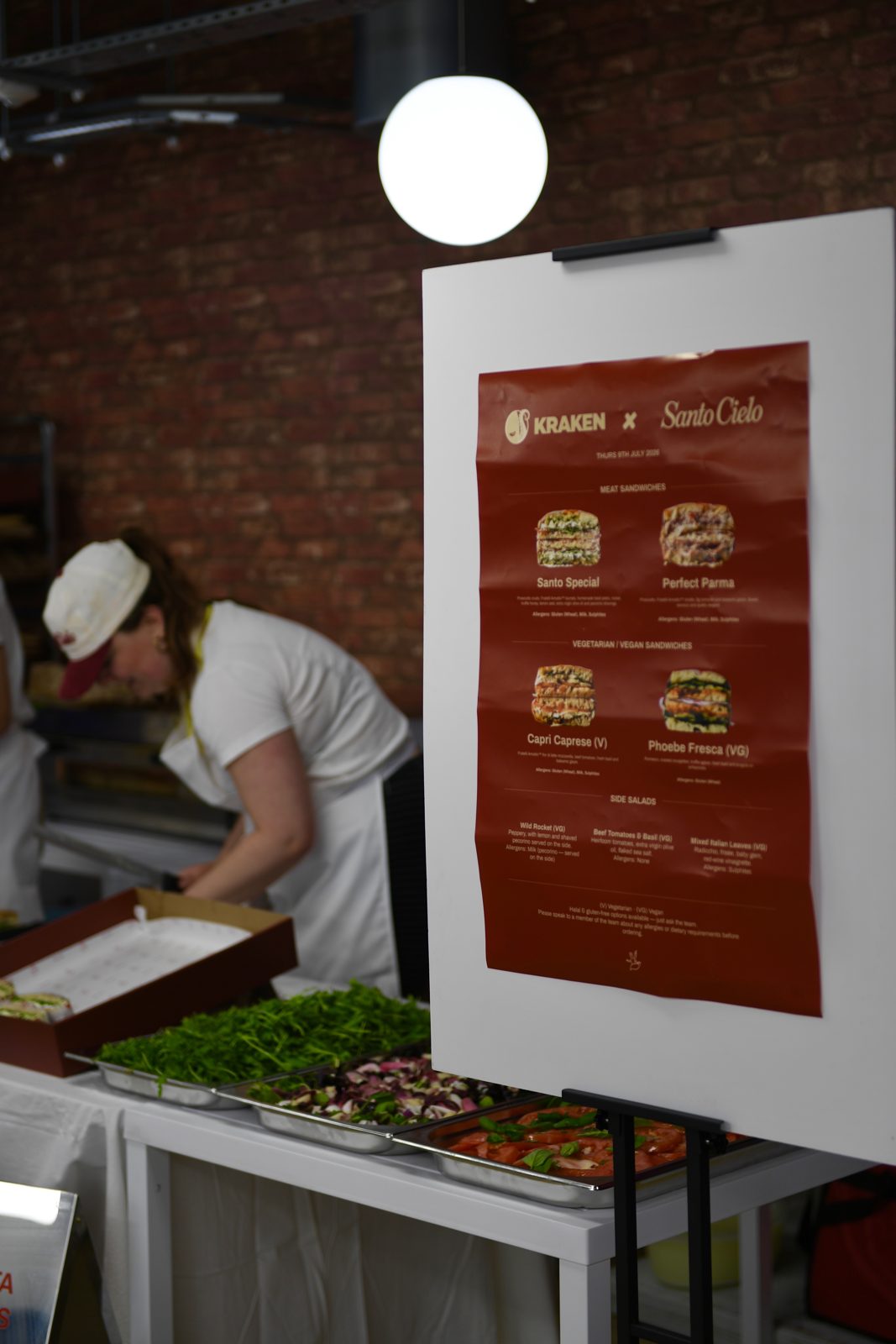 The Kraken x Santo Cielo event menu on an easel beside trays of fresh salads and platter boxes