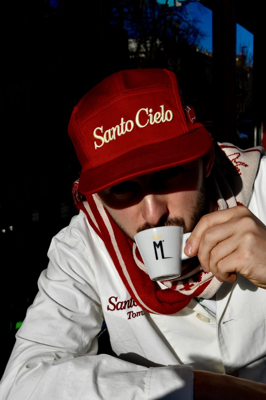 Person wearing the red Santo Cielo OG Cap with embroidered cream script logo, drinking an espresso