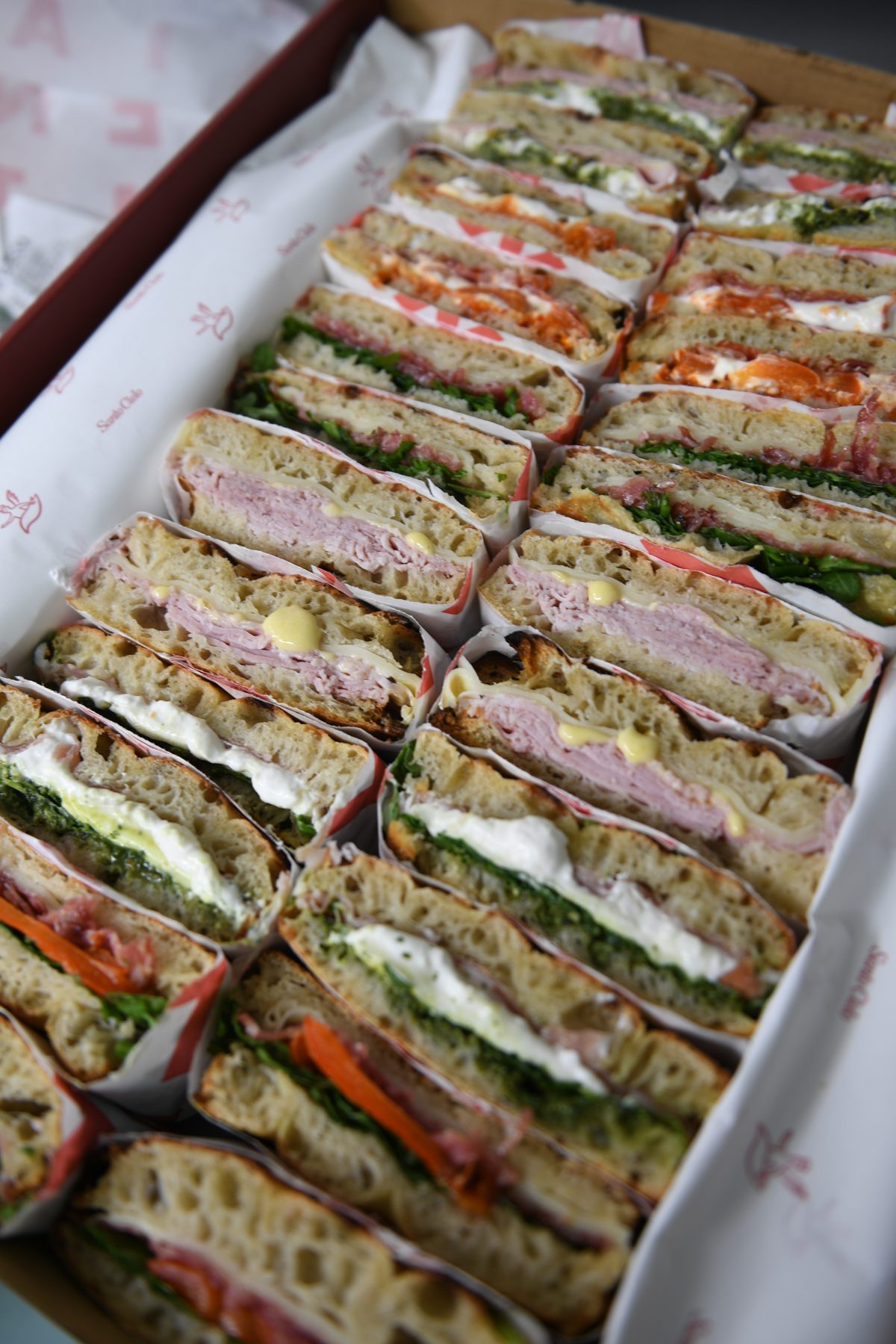 An open Santo Cielo SC Platter box packed with rows of mixed schiacciata sandwiches in branded paper