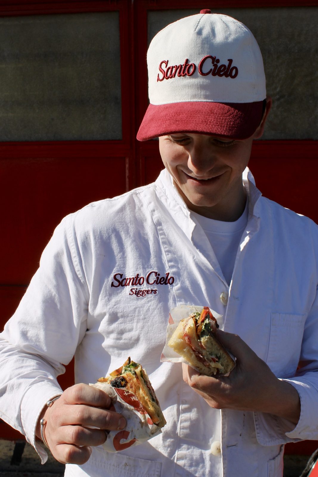 Smiling team member wearing the cream and burgundy corduroy SC Summer Cap with embroidered Santo Cielo script, holding two schiacciata halves