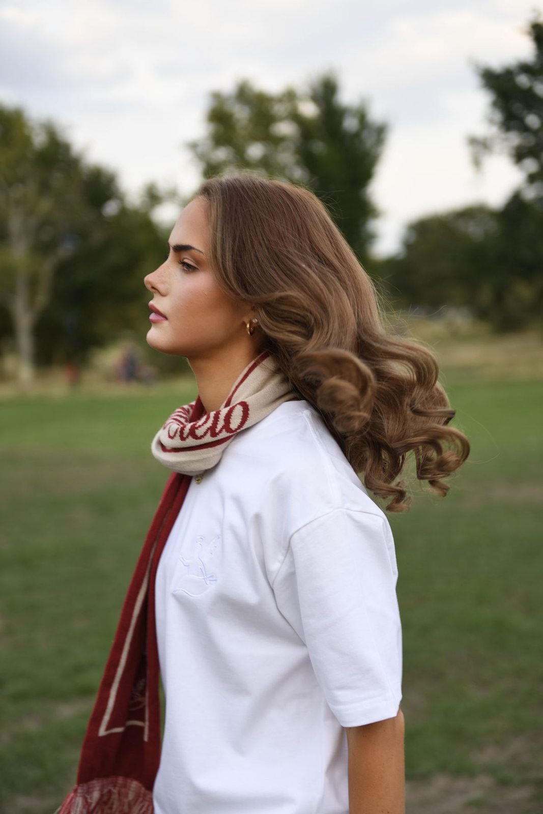 Someone in a white Santo Cielo tee with the embroidered angel and the red and cream Santo Cielo scarf, in profile in a London park