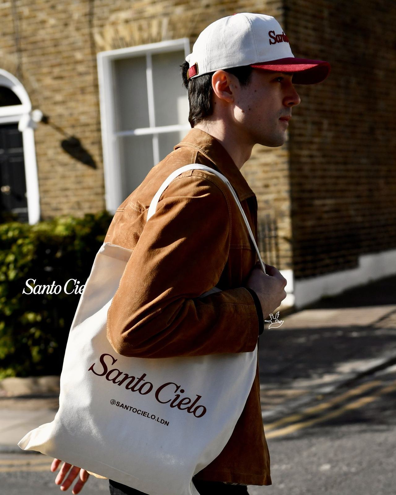 The cream Santo Cielo tote bag with the script logo and @santocielo.ldn, carried over the shoulder on a London street
