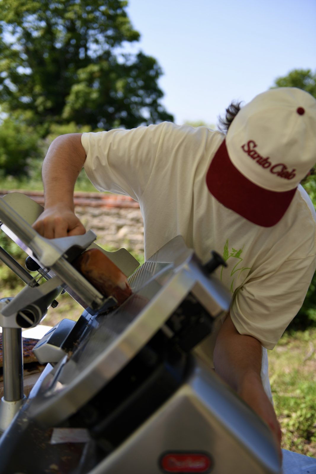 Santo Cielo team member in a branded cap slicing prosciutto on the outdoor slicer at Wildfarmed Field Day